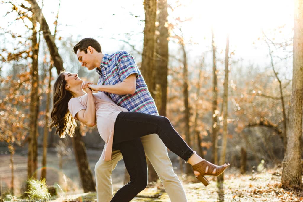 Man dips his laughing partner during a playful engagement session in golden late-day light among bare autumn trees