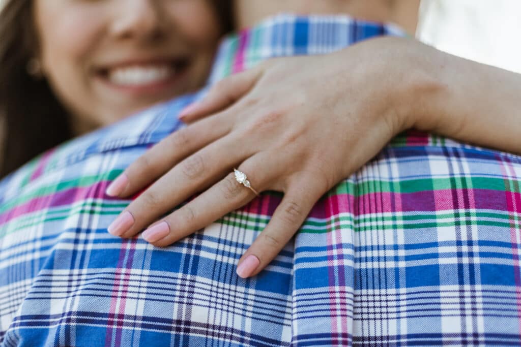 Close-up of a woman's hand with a three-stone engagement ring resting on her partner's plaid-shirted shoulder as they embrace