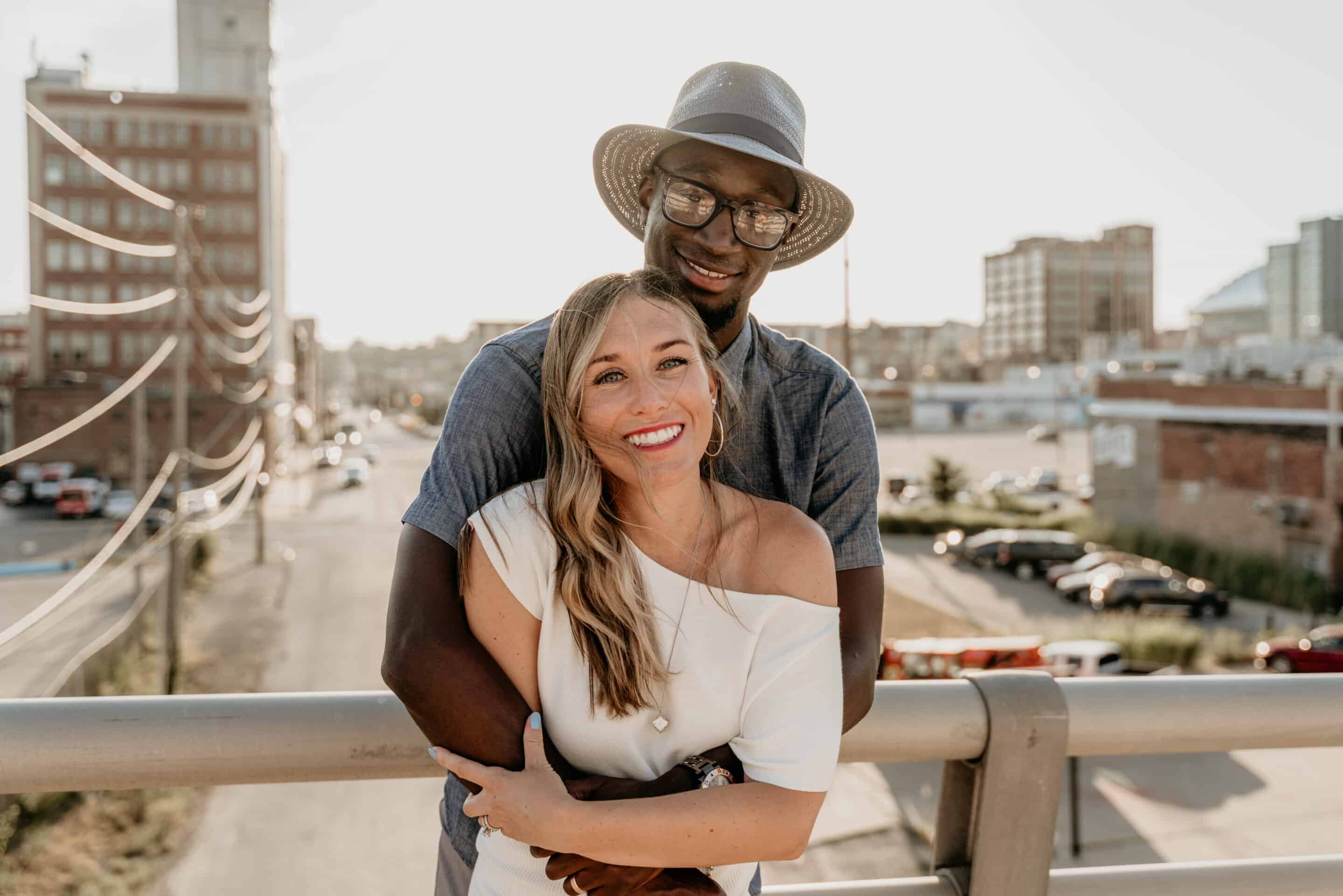 Smiling couple embrace on a bridge over the Kansas City Crossroads with string lights and the skyline glowing behind them