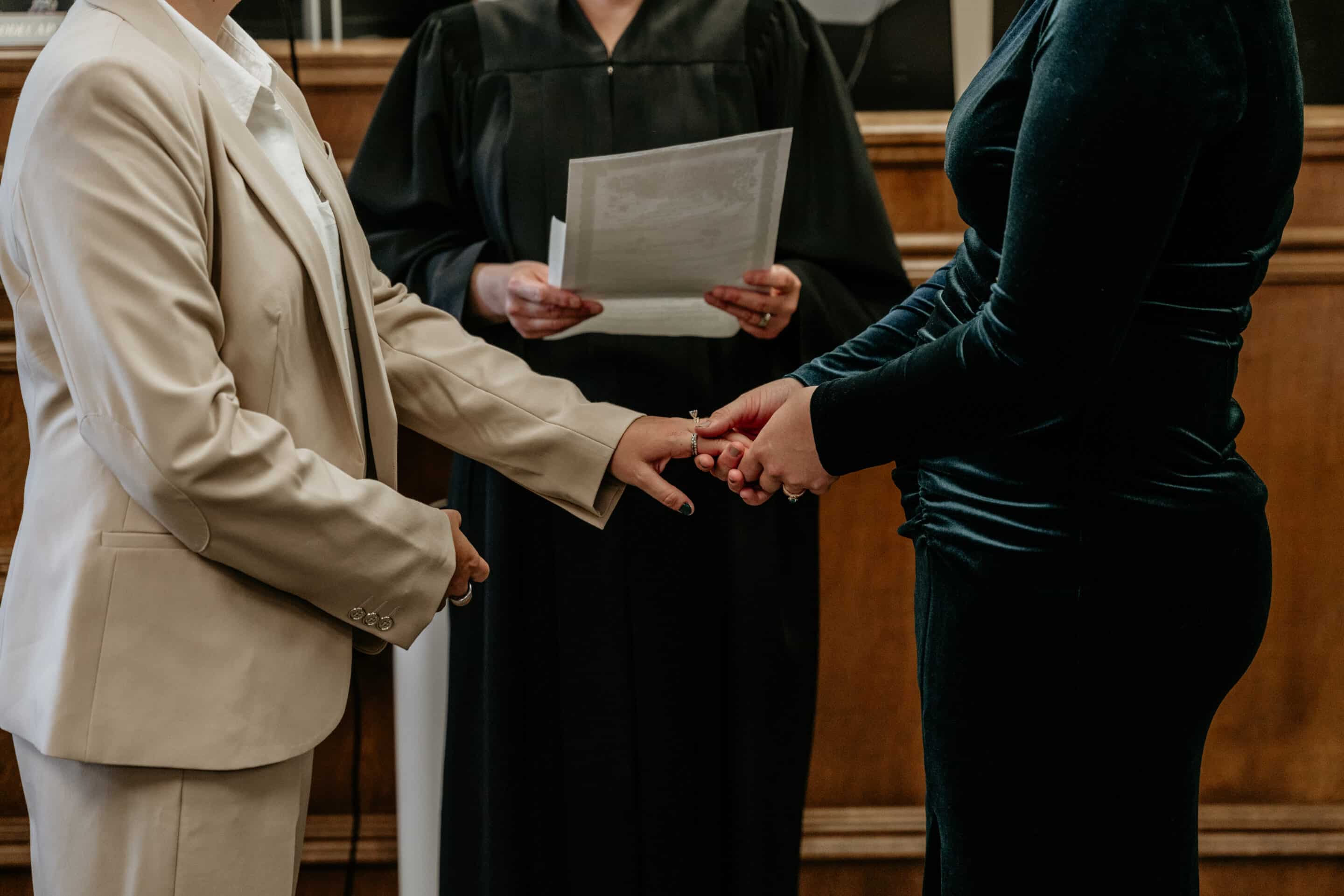 Couple holds hands before a judge in a wood-paneled courtroom during their courthouse wedding, one partner in a cream suit and the other in a dark velvet dress