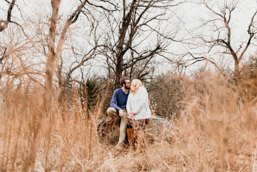 Couple kisses while seated on a rock surrounded by tall amber prairie grass and bare trees in late fall near Kansas City