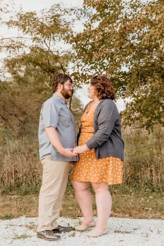 Couple holds hands under early fall trees at the edge of tall prairie grass, one partner in a mustard floral dress and gray cardigan
