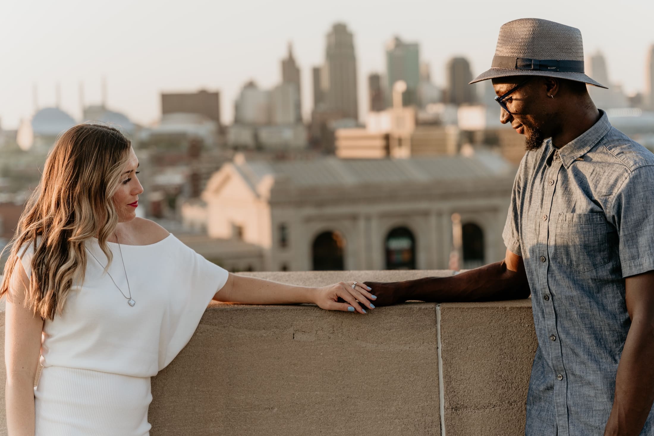 Engaged couple holds hands on the Liberty Memorial lawn at golden hour with Union Station and the downtown Kansas City skyline behind them