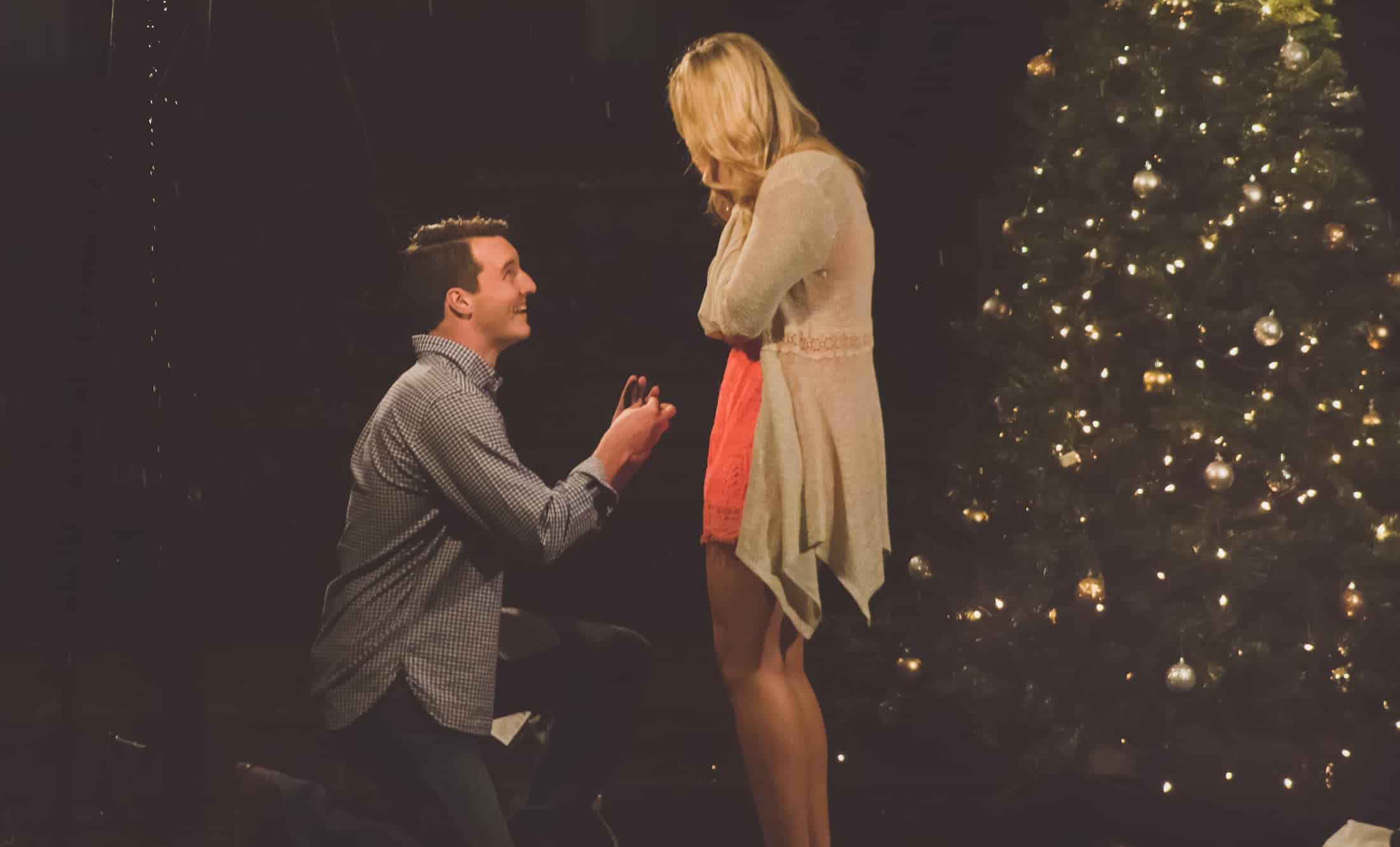 Man on one knee during a surprise proposal, holding a ring box while his partner looks down at him beside a lit Christmas tree
