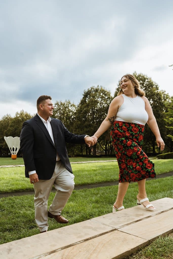 Engaged couple walks hand in hand on the Nelson-Atkins lawn in Kansas City past a Shuttlecock sculpture
