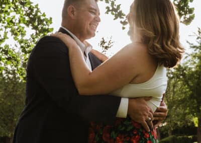 Man in a dark blazer embraces his smiling fiancee under sunlit tree branches on a grassy lawn