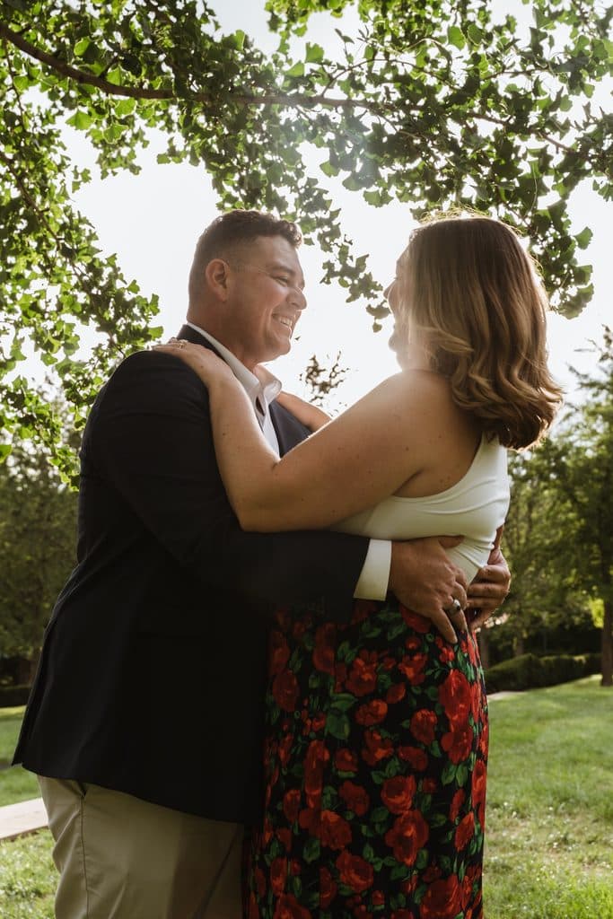 Engaged couple embraces under shade trees with her hand on his chest at the Nelson-Atkins in Kansas City
