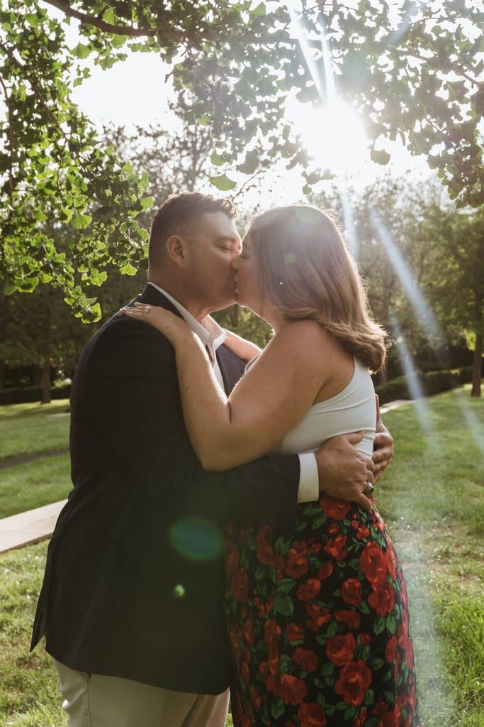 Engaged couple kisses under the trees as sunlight breaks through behind them at the Nelson-Atkins in Kansas City