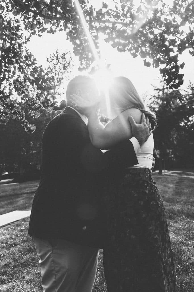 Black and white photo of an engaged couple kissing with the sun flaring through the trees at the Nelson-Atkins in Kansas City