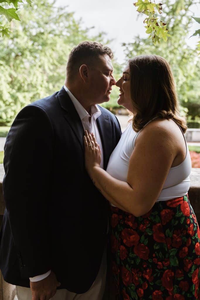 Engaged couple leans in nose to nose in front of green trees at the Nelson-Atkins in Kansas City