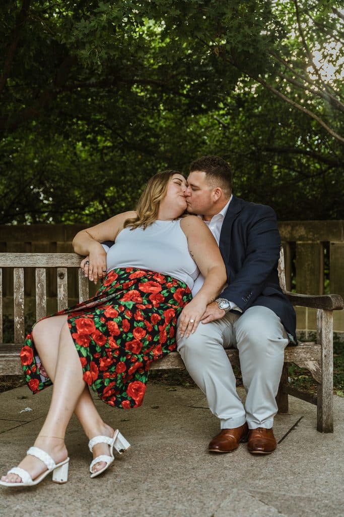 Engaged couple kisses on a wooden bench under the trees at the Nelson-Atkins in Kansas City