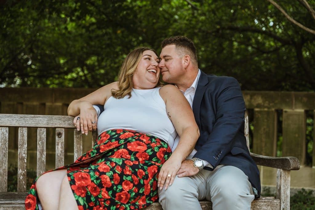 Bride-to-be laughs with her head on her fiance's shoulder on a wooden bench at the Nelson-Atkins in Kansas City
