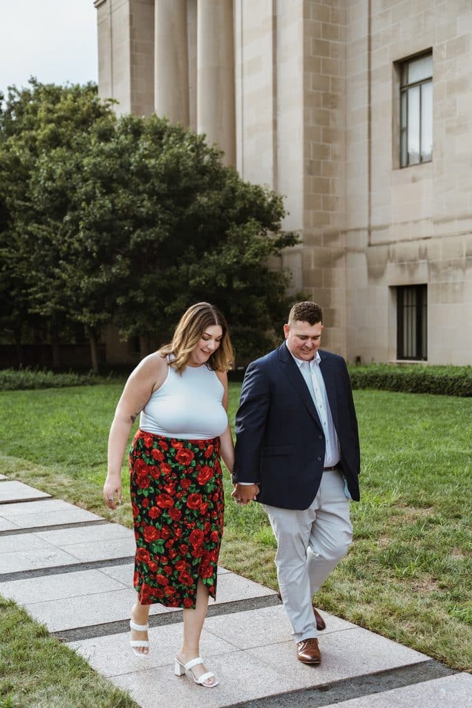 Engaged couple walks a stone path across the lawn beside the Nelson-Atkins Museum of Art building in Kansas City