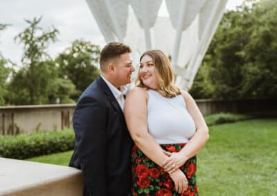 Man in a navy blazer leans in toward his smiling fiancee beside a stone wall with a Shuttlecocks sculpture behind them on the Nelson-Atkins grounds