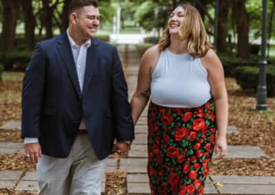 Engaged couple walk hand in hand down a stone path under green trees, the woman laughing in a white top and red floral skirt