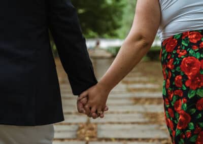View from behind of an engaged couple holding hands as they walk a leaf-scattered stone path under green trees