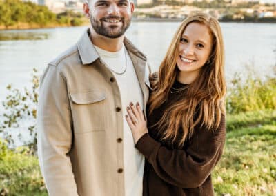 Smiling couple pose on the riverbank at Kaw Point Park, the man in a tan shirt jacket and the woman in a brown sweater, with the downtown Kansas City skyline across the water
