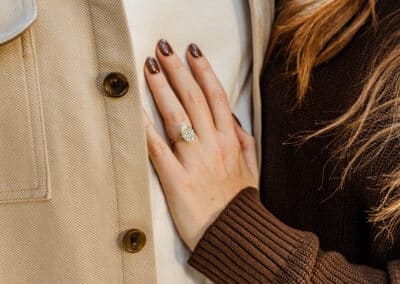 Close-up of a woman's hand with an engagement ring resting on her partner's tan shirt jacket, her long hair and brown sweater beside it