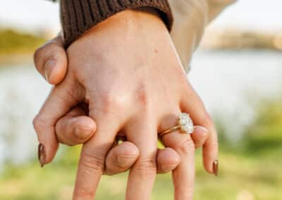 Close-up of a couple's interlaced fingers showing her engagement ring, a brown knit sleeve and green riverbank blurred behind