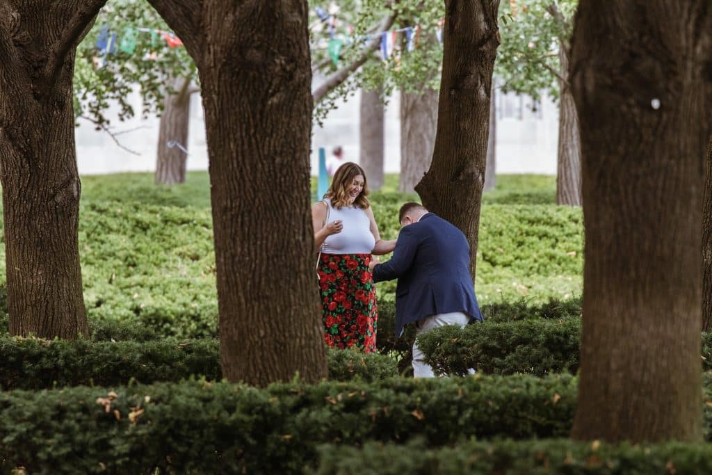 Man drops to one knee in front of his girlfriend among the hedges and linden trees at the Nelson-Atkins in Kansas City