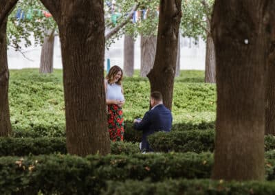 Man in a navy blazer kneels with a ring as a woman in a white top and red floral skirt looks down at him in a tree-lined garden grove