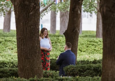 Woman in a white top and red floral skirt smiles down at her partner as he kneels on a hedge-lined lawn between large tree trunks