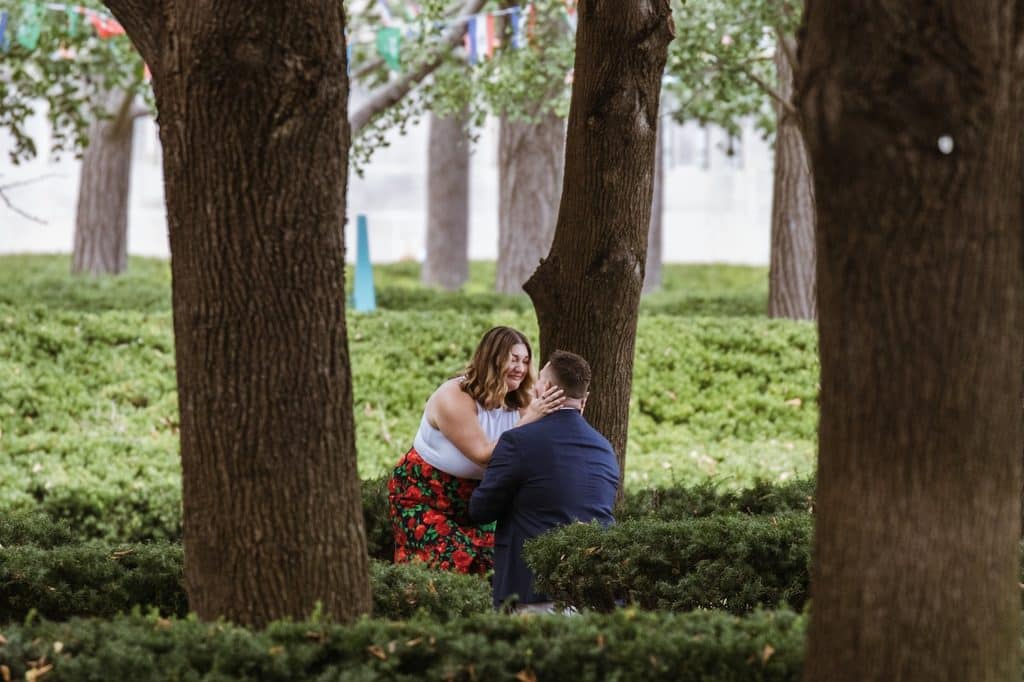 Woman wraps her arms around her kneeling fiance among the linden trees at the Nelson-Atkins in Kansas City