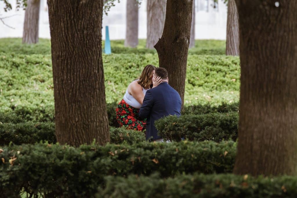 Woman holds her fiance close as he kneels on the walkway at the Nelson-Atkins in Kansas City