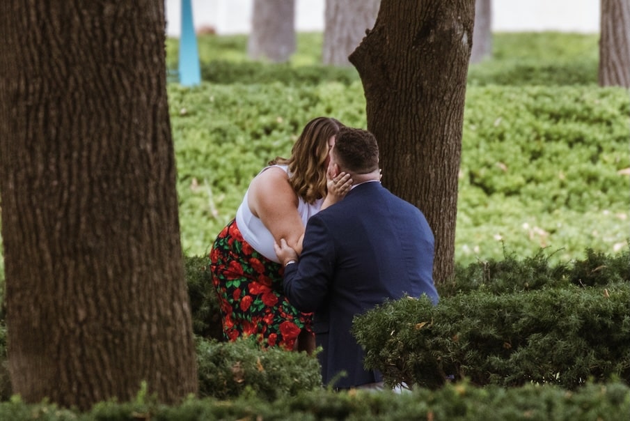 Close view of a kiss as he kneels among the trees after proposing at the Nelson-Atkins in Kansas City