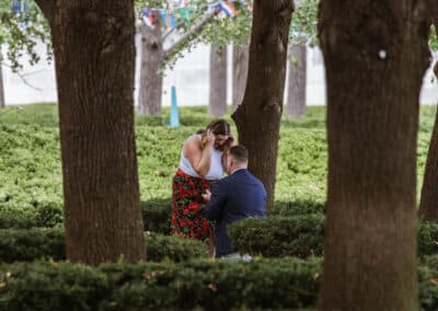 Woman covers her face with her hand in surprise as her partner kneels before her among hedges and tall trees