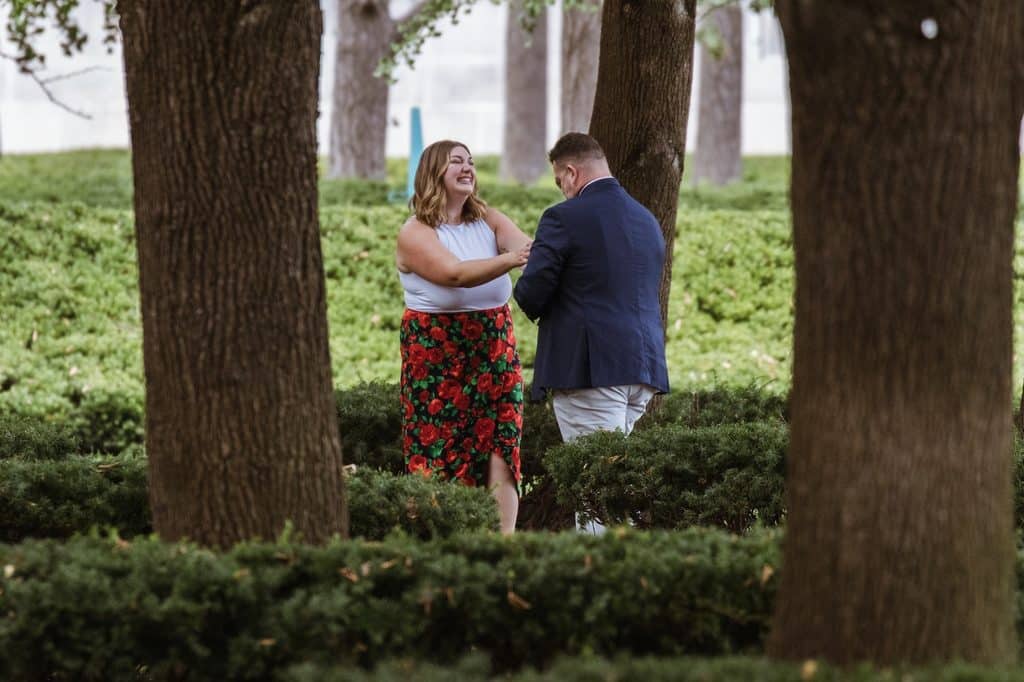 Bride-to-be laughs with her hands together as her fiance turns toward her among the linden trees at the Nelson-Atkins in Kansas City