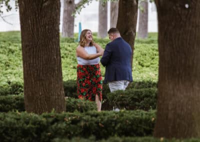 Newly engaged couple stand face to face and smile in a hedge-lined garden grove right after the proposal
