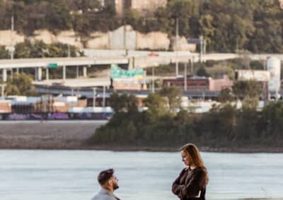 Man kneels to propose on a grassy riverbank at Kaw Point Park with the downtown Kansas City skyline, including City Hall, across the water
