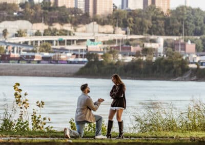 Man on one knee holds out a ring to a woman in a brown sweater and tall black boots at Kaw Point Park, the downtown Kansas City skyline across the river