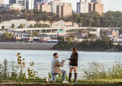 Woman in a brown sweater reaches for her partner's hand as he kneels with a ring at Kaw Point Park, the Kansas City skyline across the river